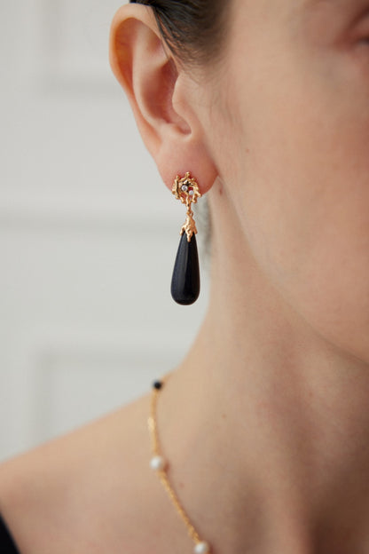 Close-up of a person wearing Starry Night Blue Sandstone Drop Earrings in vintage gold by Siren Mode, featuring long black teardrop-shaped pendants. Part of a gold chain necklace with pearls is partially visible. The background is softly blurred in neutral tones.