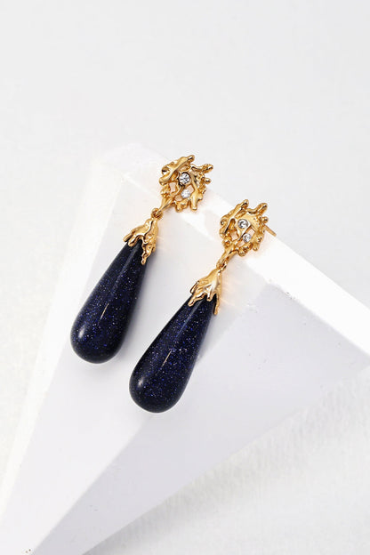 The Starry Night Blue Sandstone Drop Earrings - Vintage Gold by Siren Mode exude vintage charm with teardrop-shaped blue sandstone stones and small crystals near the top, all supported by an intricate gold design. Displayed on a white geometric stand, these captivating 925 silver treasures are truly mesmerizing.