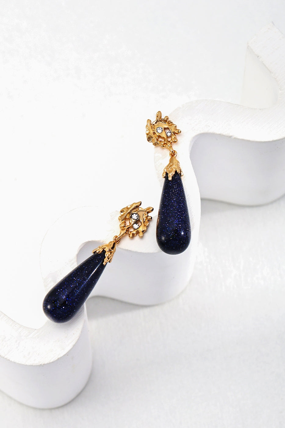 The Starry Night Blue Sandstone Drop Earrings by Siren Mode feature deep blue sandstone stones elegantly set in vintage gold, adorned with small white crystals at the top. These charming earrings are displayed on a white, wavy stand.