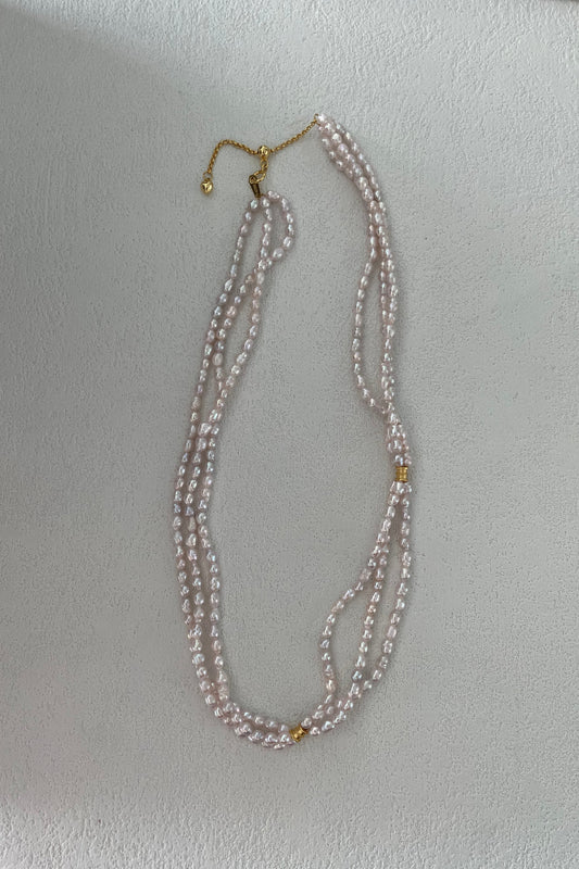 The Siren Mode Triple Strand Pearl Necklace, crafted in sterling silver with 18K gold plating, showcases small freshwater pearls and features a gold clasp along with two gold beads interspersed among the pearls. The necklace is elegantly laid out flat on a light-colored surface.