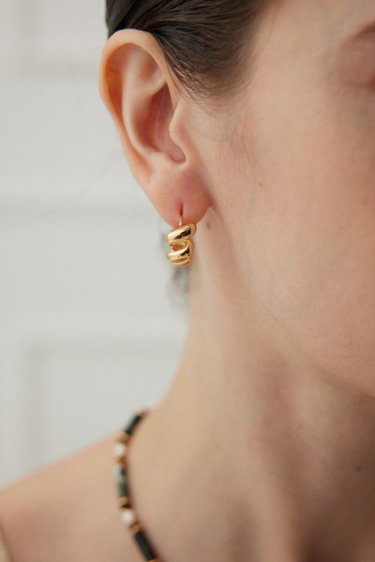 Close-up of a woman's ear adorned with Siren Mode's Twisted Harmony Hoop Earrings in vintage gold. Her straight brown hair is pulled back, revealing part of a multicolored beaded necklace. The softly lit and out-of-focus background accentuates the versatile elegance of these hoop earrings.