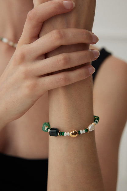 A close-up of a person wearing the "Verdant Treasure Bracelet - Vintage Gold with Natural Stones and Pearls" by Siren Mode. The bracelet features green, white, and black beads, along with a small gold charm. Delicate 925 silver accents are also included. The person is dressed in a black sleeveless top and is holding their wrist with their other hand, showcasing this stylish accessory.