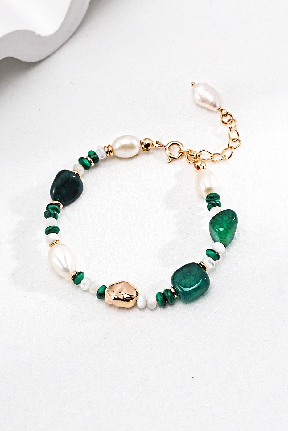 The Verdant Treasure Bracelet by Siren Mode features green stones, irregular pearls, and vintage gold-toned accents. It is secured with a 925 silver clasp and is displayed on a white surface, with shadows adding depth to the image.