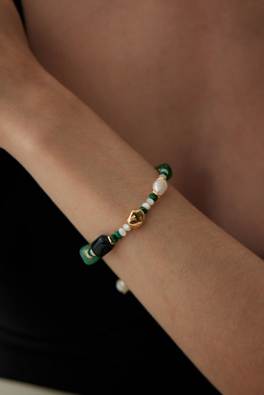 Close-up of a woman's arm adorned with the Verdant Treasure Bracelet by Siren Mode. The adjustable bracelet features a combination of green, white, and black natural stones and pearls with a central vintage gold bead. The woman is wearing a black top, and her light skin accentuates the jewelry's elegance.