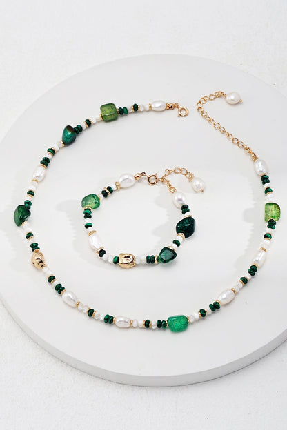 A jewelry set from Siren Mode, featuring the Verdant Treasure Bracelet in vintage gold with natural stones and pearls, lies on a round white surface. This exquisite set includes a necklace and an adjustable bracelet, both adorned with green beads, irregular pearls, and small golden accents, complemented by 925 silver clasps. The necklace is elegantly arranged in a circular shape with the bracelet nestled inside it.