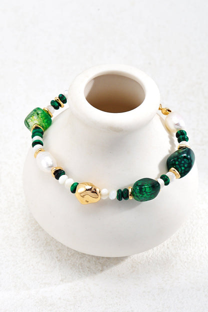 A stylish Verdant Treasure Bracelet by Siren Mode, featuring green and white beads with vintage gold accents, is elegantly draped over a small white vase. This adjustable bracelet includes various shapes and sizes of natural stones and pearls, with prominent green stones, 925 silver details, and a small gold charm, creating an elegant appearance.