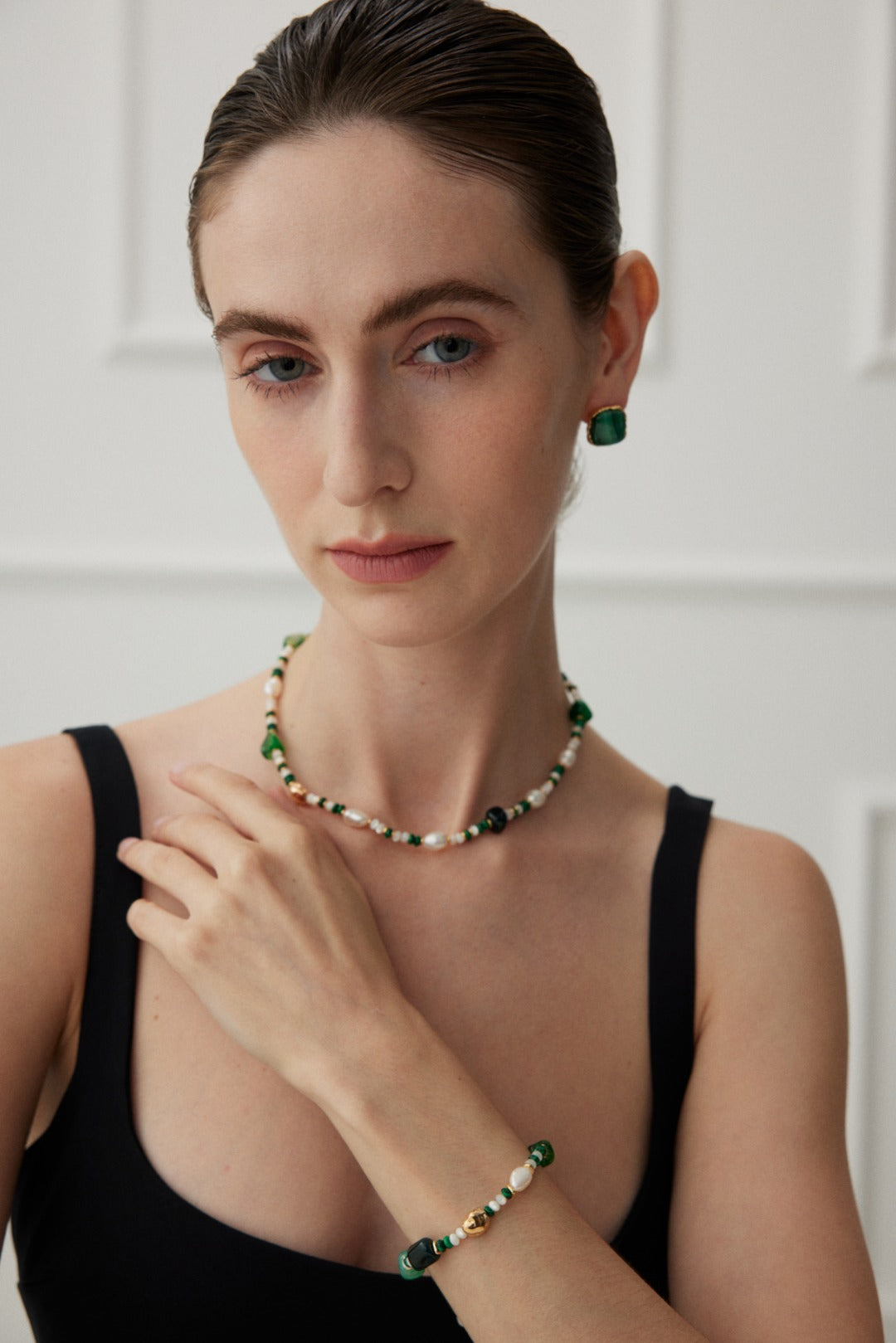 A woman with slicked-back hair wears a black sleeveless top, a beaded necklace of irregular pearls, matching earrings, and the Verdant Treasure Bracelet from Siren Mode. The beads are green and white, with gold accents on 925 silver. She gazes softly at the camera with one hand touching her neck against the softly lit room background.