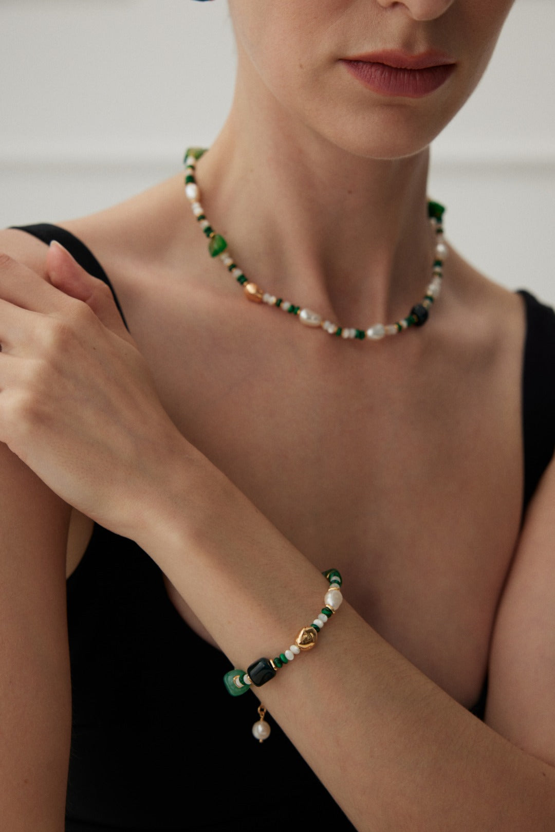 A woman wearing a beaded necklace, as well as the Verdant Treasure Bracelet by Siren Mode—an adjustable bracelet adorned with green, white, and gold beads, alongside irregular pearls. She is dressed in a sleeveless black top and has a neutral expression, with her arms crossed over her chest. Only her lower face is visible.