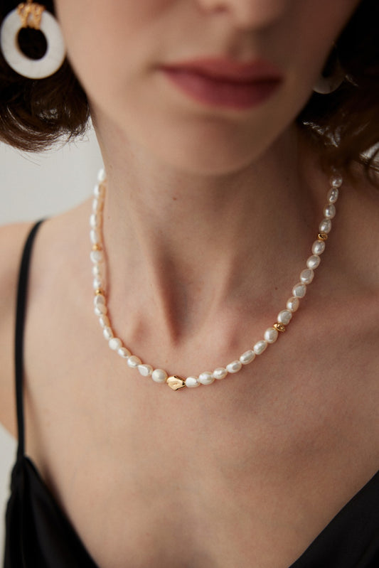 A close-up captures a woman wearing the Siren Mode Vintage Gold 925 Silver Baroque Pearl Necklace. She is dressed in a black sleeveless top and pairs the necklace with circular white earrings. The blurred background draws attention to the exquisite jewelry.