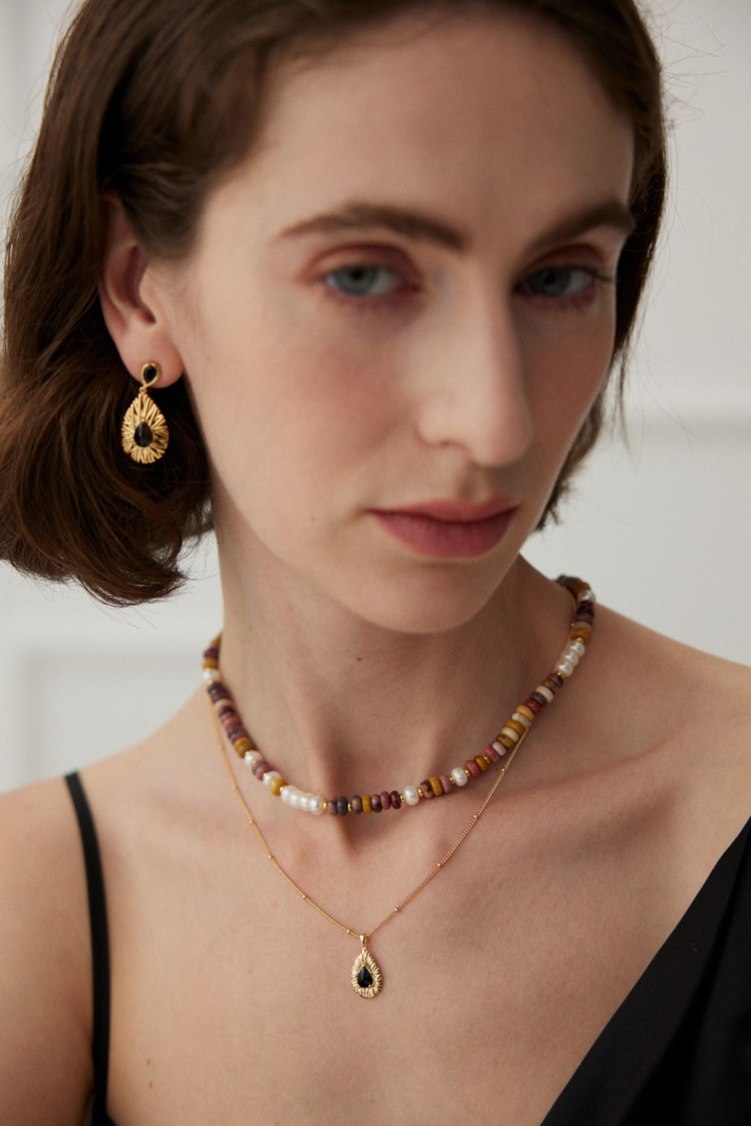 Close-up of a person with short brown hair, wearing a black top. They have a serious expression and are adorned with Siren Mode's Vintage Gold 925 Silver Black Zircon Drop Earrings, a beaded necklace, and a delicate gold necklace with a teardrop-shaped pendant. The background is a softly lit white room.