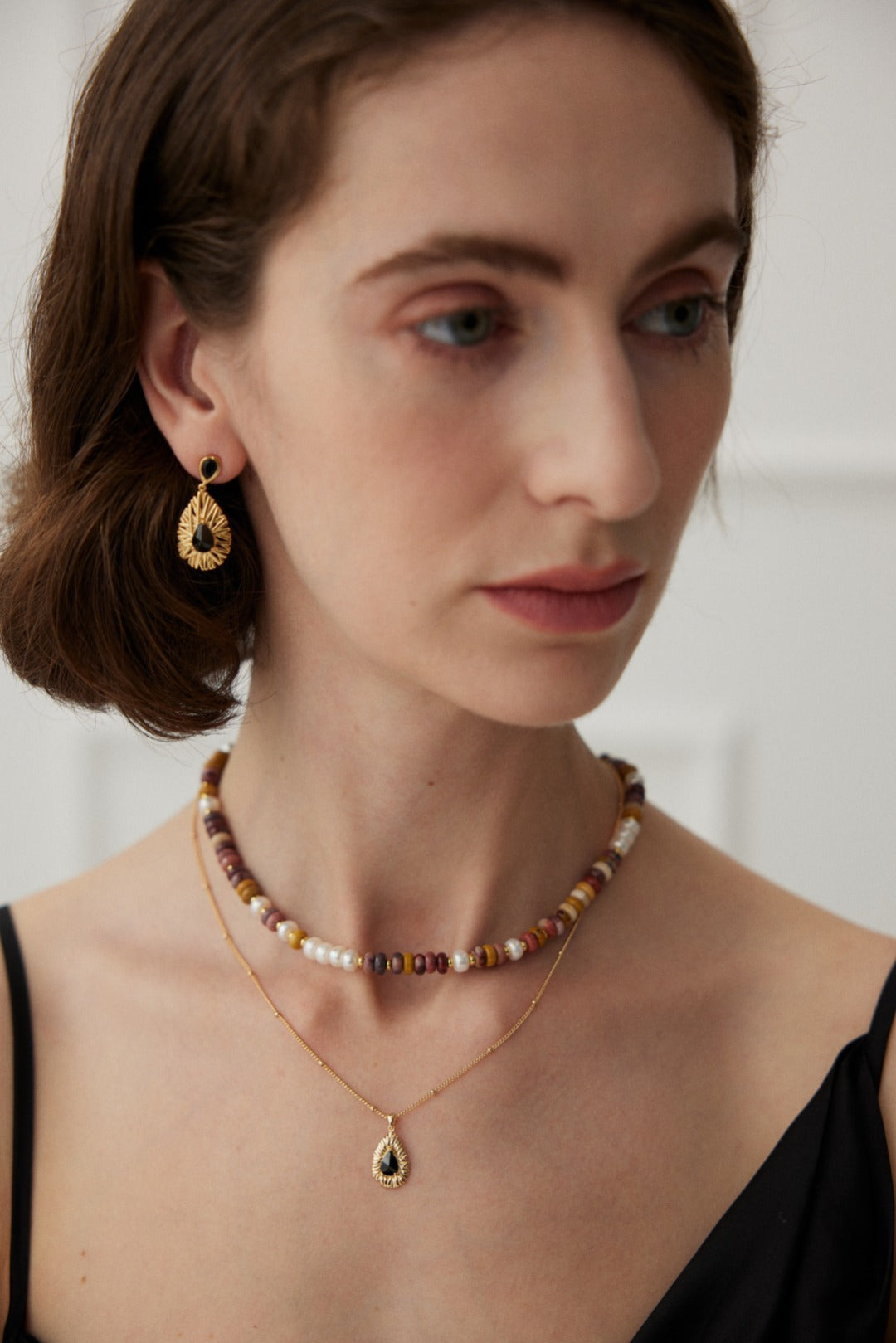 A person with short brown hair is shown wearing a black dress. They are adorned with Siren Mode's Vintage Gold 925 Silver Black Zircon Drop Earrings, a beaded choker necklace with multicolored beads, and a delicate gold chain necklace featuring a teardrop-shaped pendant. The background is softly blurred and neutral-toned.