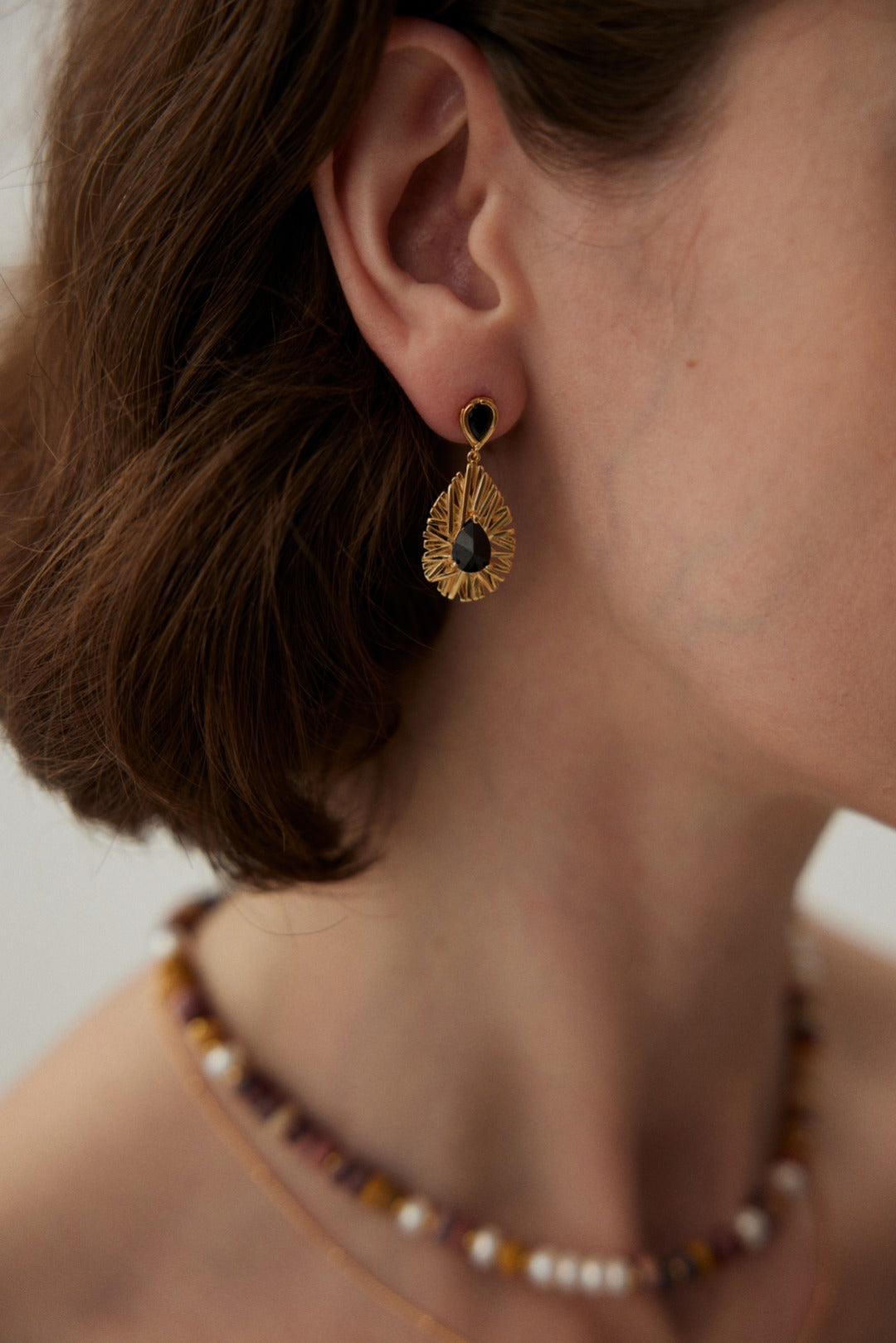 Close-up of a woman with short wavy brown hair wearing Siren Mode's Vintage Gold 925 Silver Black Zircon Drop Earrings, featuring a unique and intricate design. She is also adorned with a beaded necklace in varying colors. The background is blurred to emphasize her elegant jewelry.