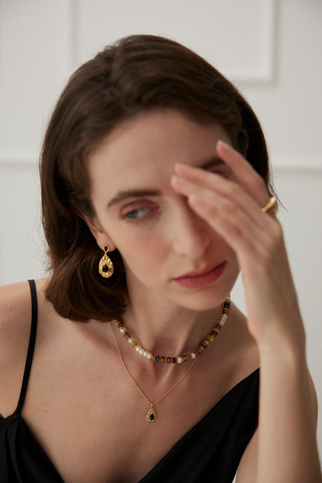 A woman with shoulder-length brown hair is wearing a black sleeveless top, a beaded necklace, a gold chain with a pendant, and Siren Mode's Vintage Gold 925 Silver Black Zircon Drop Earrings. She also sports a gold ring. She is touching her forehead with her right hand, and her expression appears thoughtful. The background is white and minimalistic.