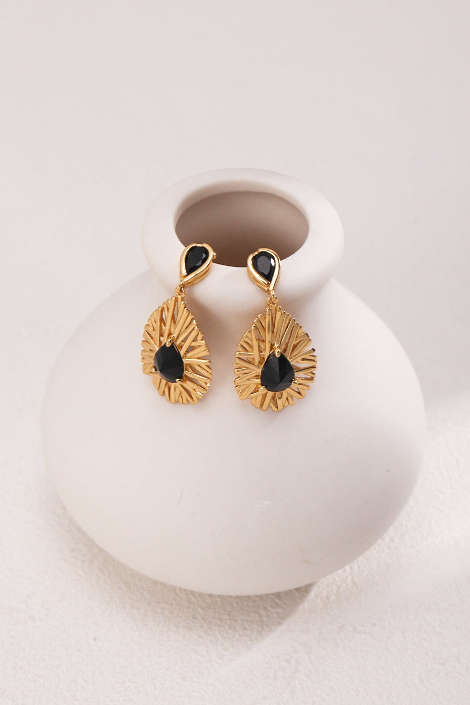 A pair of Vintage Gold 925 Silver Black Zircon Drop Earrings from Siren Mode hang on the rim of a small, round, white ceramic vase with a narrow opening. The textured design around the stones creates a radiant pattern. These stunning black zircon earrings rest on a light surface, exuding elegance.