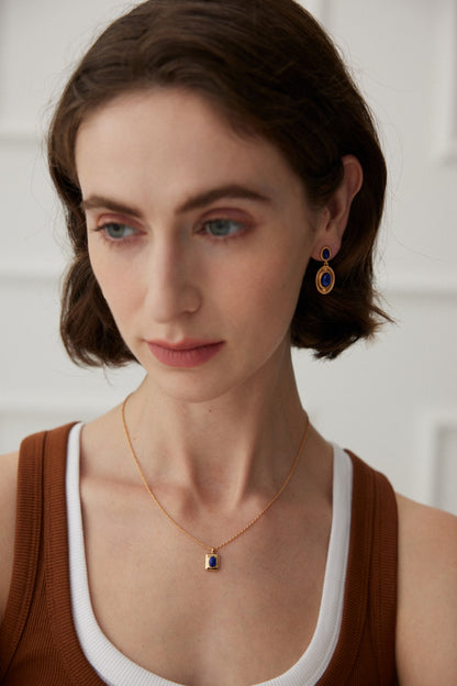 A person with short brown hair and fair skin is shown wearing a brown tank top with a white undershirt. They are accessorized with Siren Mode's Vintage Gold 925 Silver Lapis Lazuli Drop Earrings and a matching gold necklace with a blue pendant. The background is a soft white.