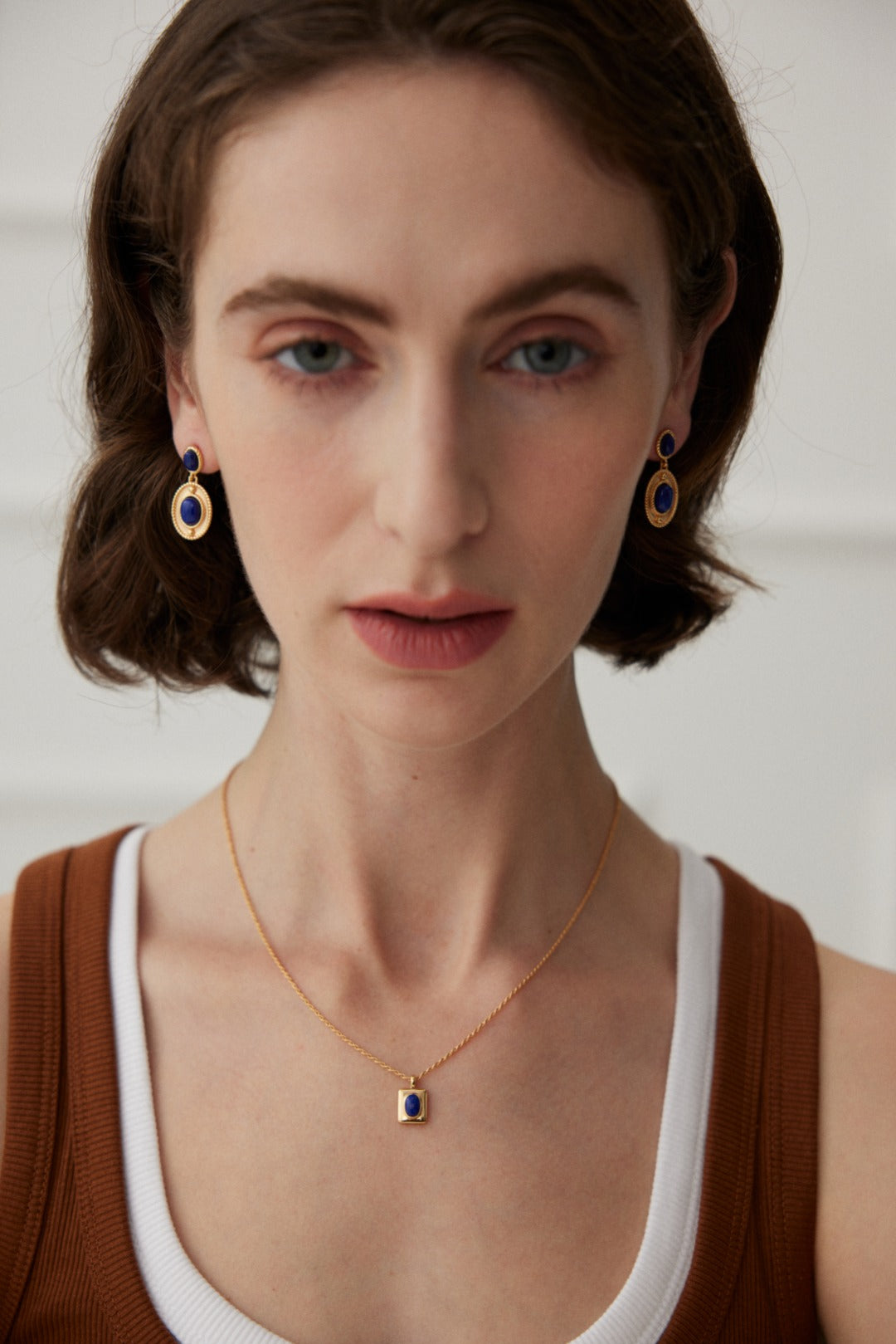 A woman with short brunette hair is wearing a sleeveless brown top over a white tank top. She has Siren Mode's Vintage Gold 925 Silver Lapis Lazuli Drop Earrings and a matching necklace. The background is plain and light-colored.