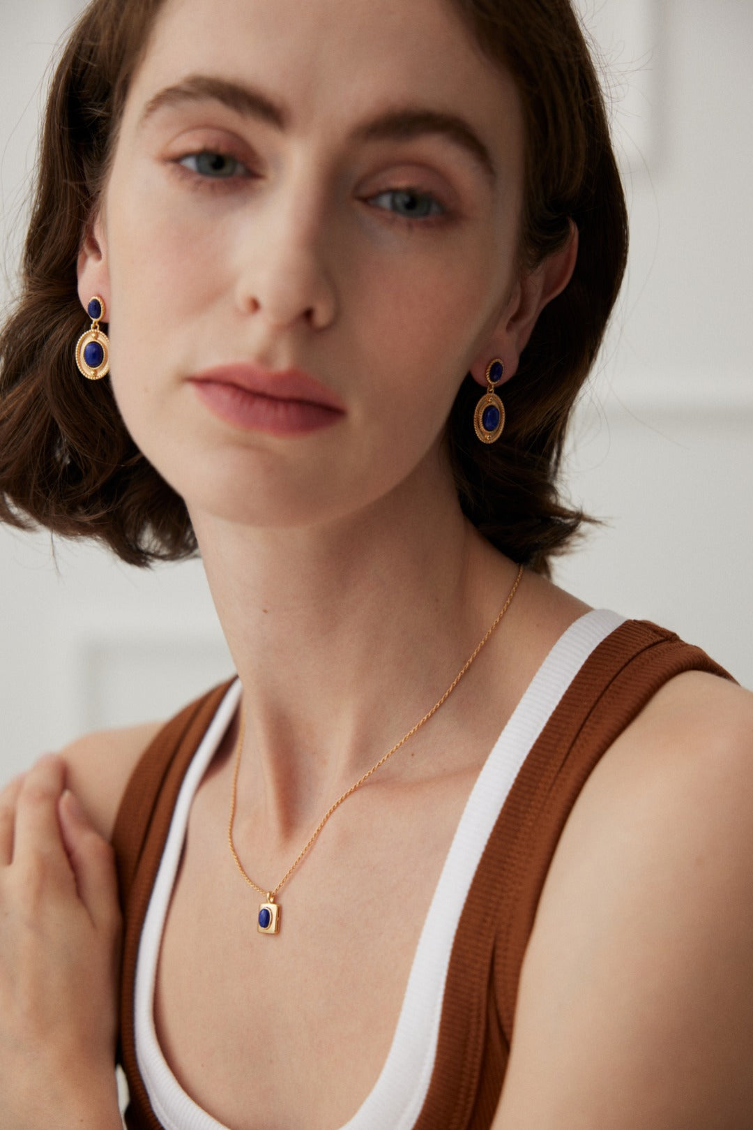 A person with short brown hair is wearing a white top underneath a brown sleeveless garment. They are adorned with matching jewelry, featuring Siren Mode's Vintage Gold 925 Silver Lapis Lazuli Drop Earrings and a 925 silver necklace with a blue oval pendant. They appear serene and are looking forward.