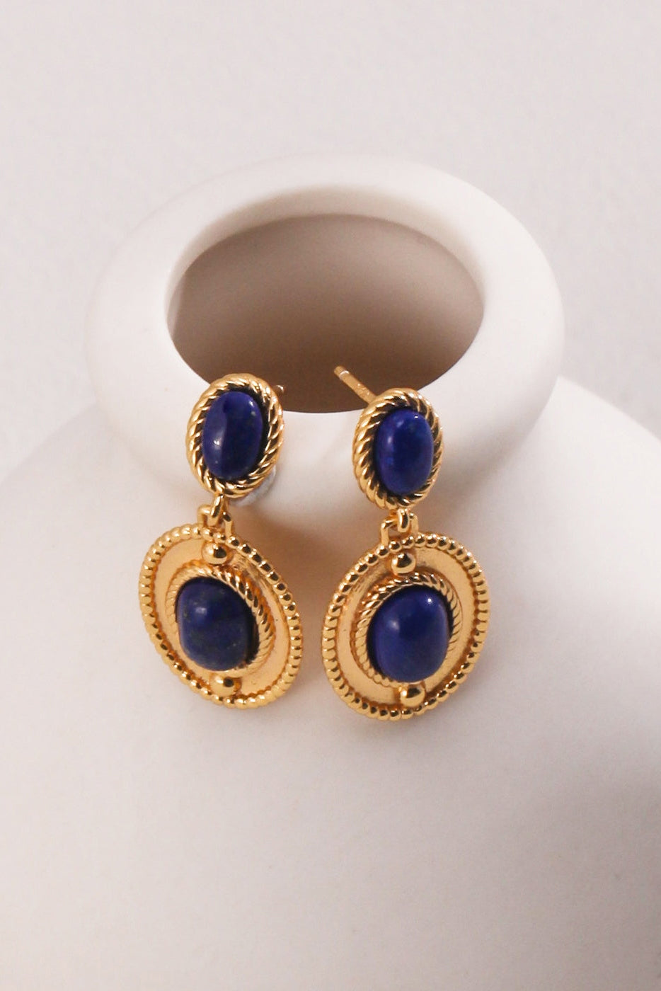 A pair of elegant Vintage Gold 925 Silver Lapis Lazuli Drop Earrings from Siren Mode, featuring oval blue gemstones. The top part of each earring showcases a smaller stone, while the larger lapis lazuli gemstone is set in an intricately designed circular gold frame below. These stunning earrings are displayed on a white vase.