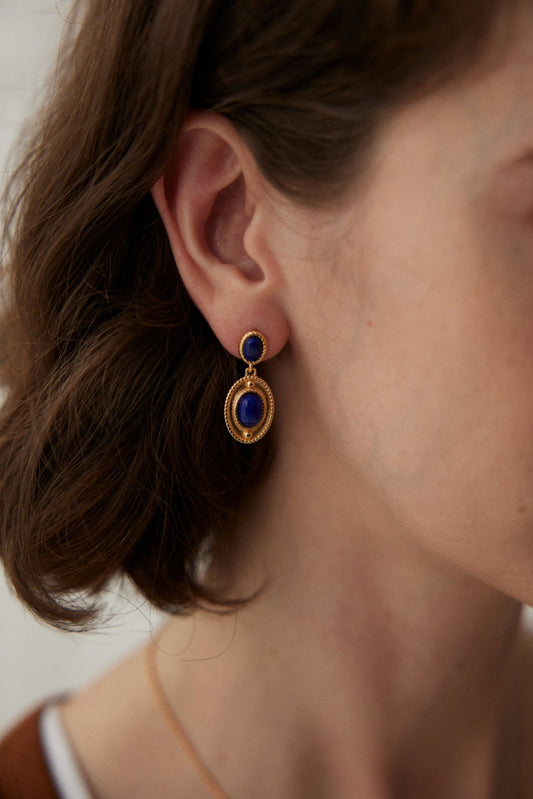 Close-up of a person wearing the Vintage Gold 925 Silver Lapis Lazuli Drop Earrings by Siren Mode. The elegant earring features two circular lapis lazuli stones; the smaller one is attached to the earlobe, while the larger one hangs within a gold frame just below. Brown hair partially covers the ear, enhancing its vintage allure.