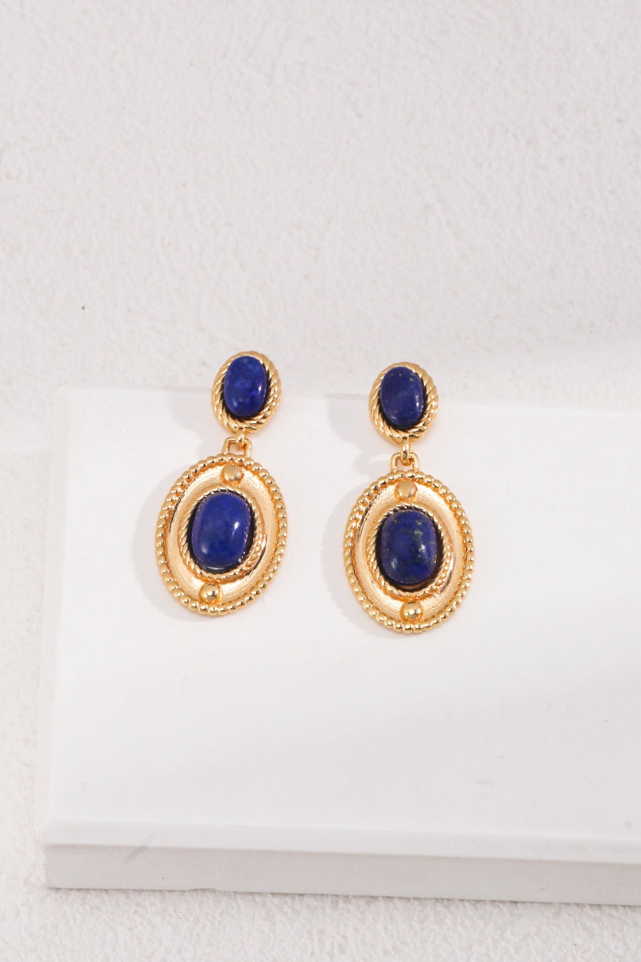 A pair of Siren Mode's Vintage Gold 925 Silver Lapis Lazuli Drop Earrings. Each earring features a small blue stone set in gold at the top and a larger lapis lazuli stone encircled by a gold frame suspended below. They are displayed on a white surface.