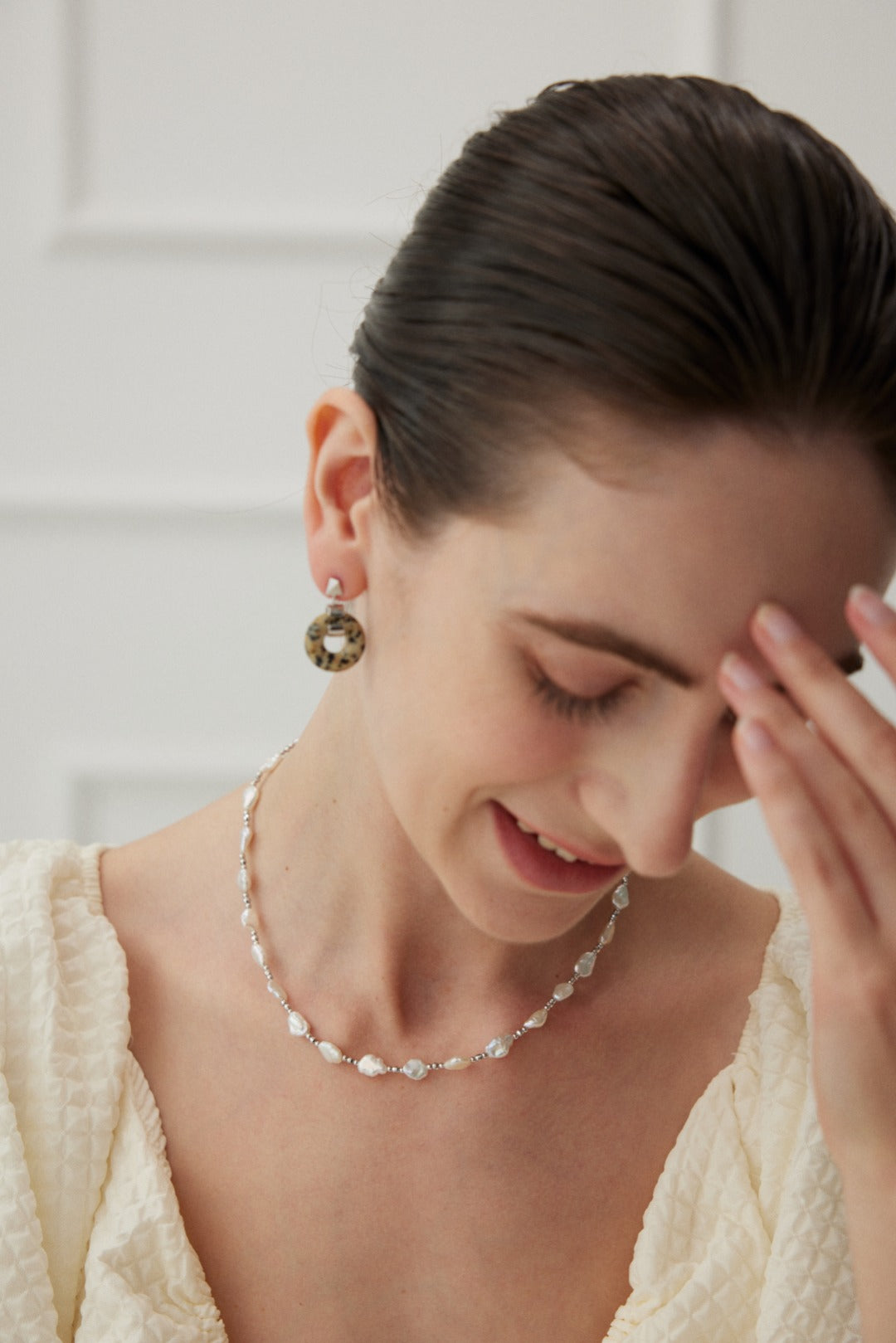 A woman with dark hair slicked back is smiling and looking down. She is wearing a pearl necklace, Siren Mode's Vintage Gold 925 Silver Leopard Stone and Tiger's Eye Earrings, and a light-colored textured top with a low neckline. Her hand, adorned with a ring featuring Tiger's Eye set in 925 silver, gently touches her forehead. The background shows a white wall with molding details.