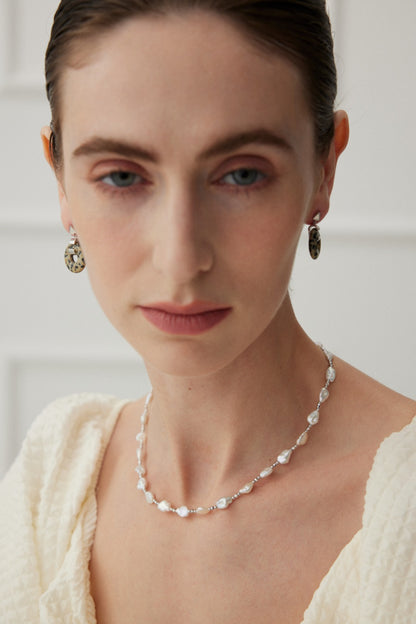 A woman with light skin, slicked-back hair, and minimal makeup wears Siren Mode's Vintage Gold 925 Silver Leopard Stone and Tiger's Eye Earrings and a delicate pearl necklace. She is dressed in a textured white garment and gazes softly forward with a neutral expression, set against a neutral, slightly blurred background.