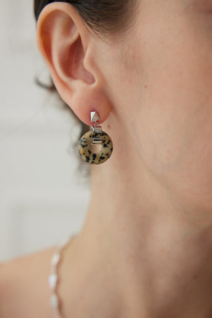 Close-up of a person wearing Siren Mode's Vintage Gold 925 Silver Leopard Stone and Tiger's Eye Earrings. The earring features a round, speckled Leopard Stone hanging from a small geometric 925 Silver stud with a light brown and black spotted pattern. The background is neutral, and part of the person's ear, neck, and shoulder are visible.