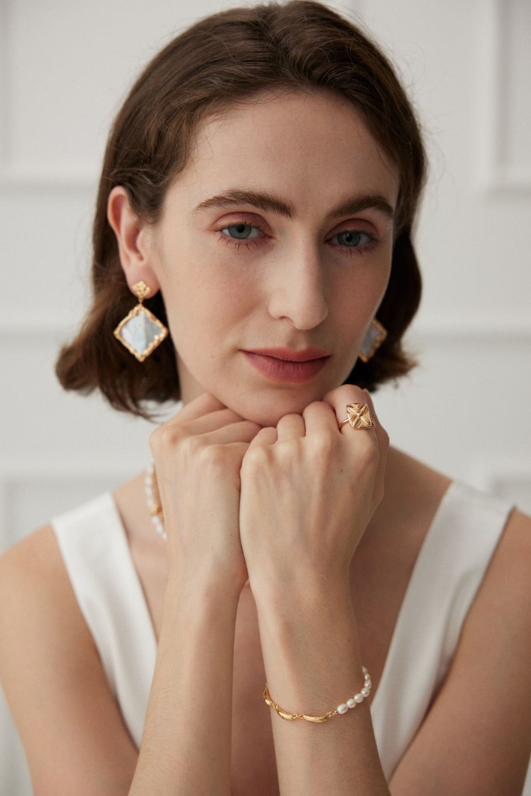 A woman with short brown hair wears gold and natural pearl jewelry, including geometric earrings, a Vintage Gold 925 Silver Natural Pearl Bracelet by Siren Mode, and a ring. She rests her chin on her hands, gazing into the camera with a serene expression. She is dressed in a white sleeveless top.