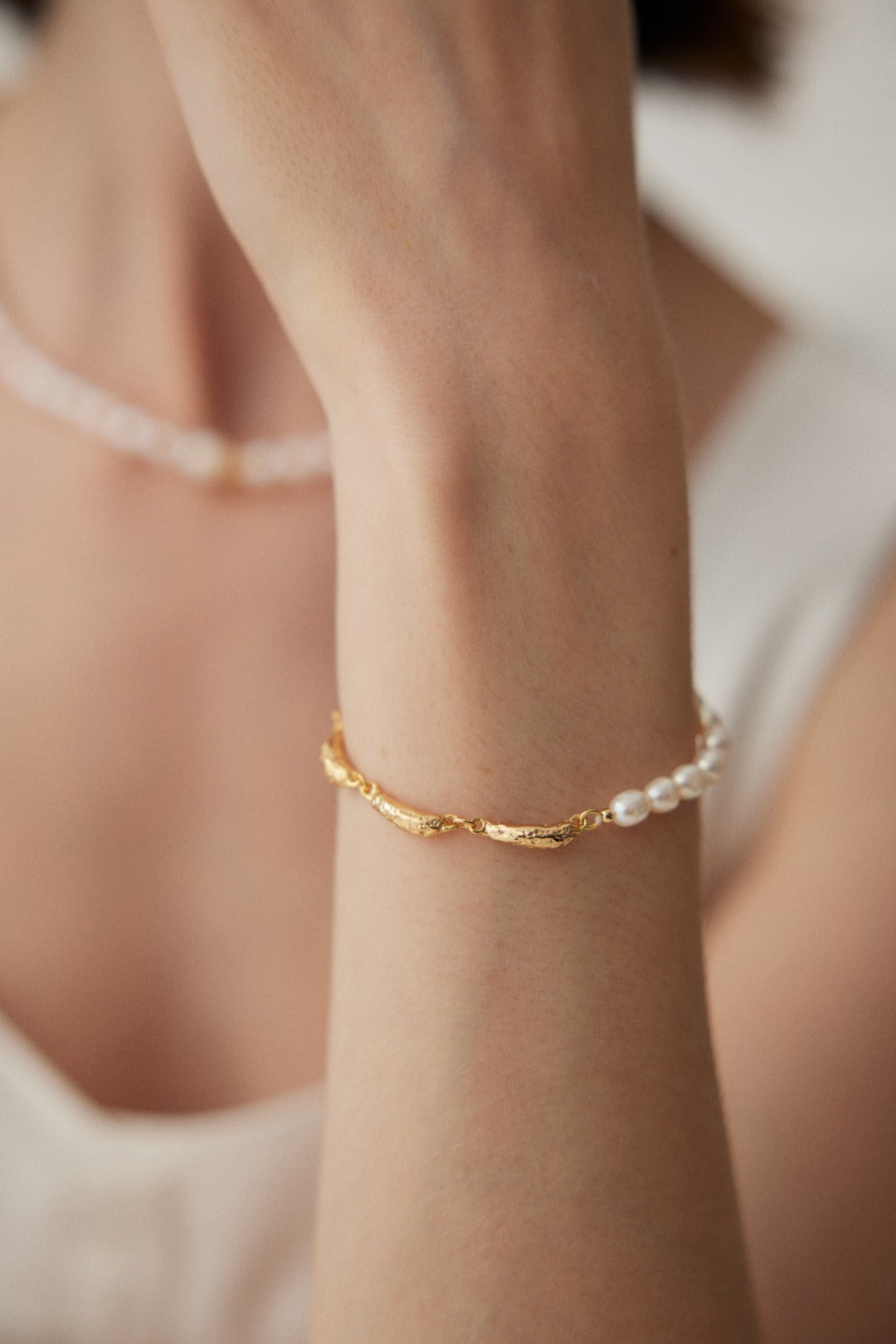 Close-up of a person's wrist wearing a Siren Mode Vintage Gold 925 Silver Natural Pearl Bracelet. The person is also seen wearing a matching pearl necklace. The background is softly blurred, and the person is dressed in a white top.