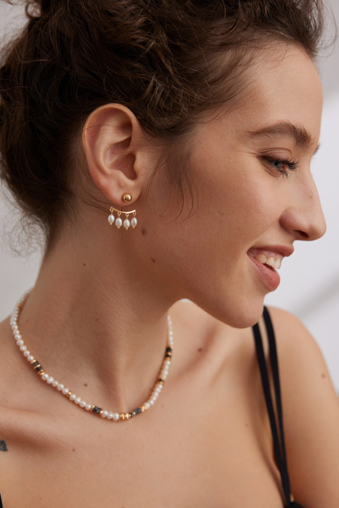 A woman with brown hair styled in an updo is smiling and looking to the side. She is wearing Siren Mode Vintage Gold 925 Silver Natural Pearl Drop Earrings and a matching pearl necklace. She has a light complexion and is dressed in a black spaghetti strap top.