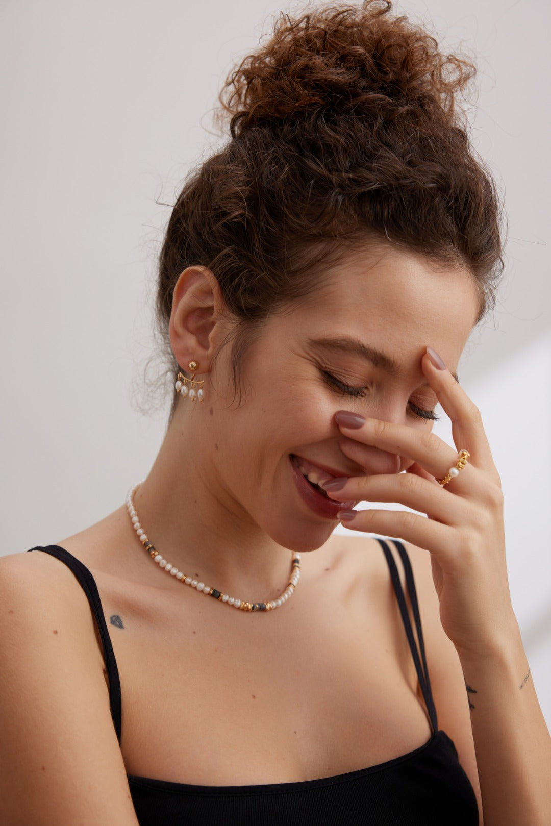 A woman with curly hair in a bun is smiling and looking down while touching her forehead with one hand. She is wearing a black tank top, Siren Mode's Vintage Gold 925 Silver Natural Pearl Drop Earrings, a pearl necklace, and a gold ring. She has a small heart tattoo on her upper arm.