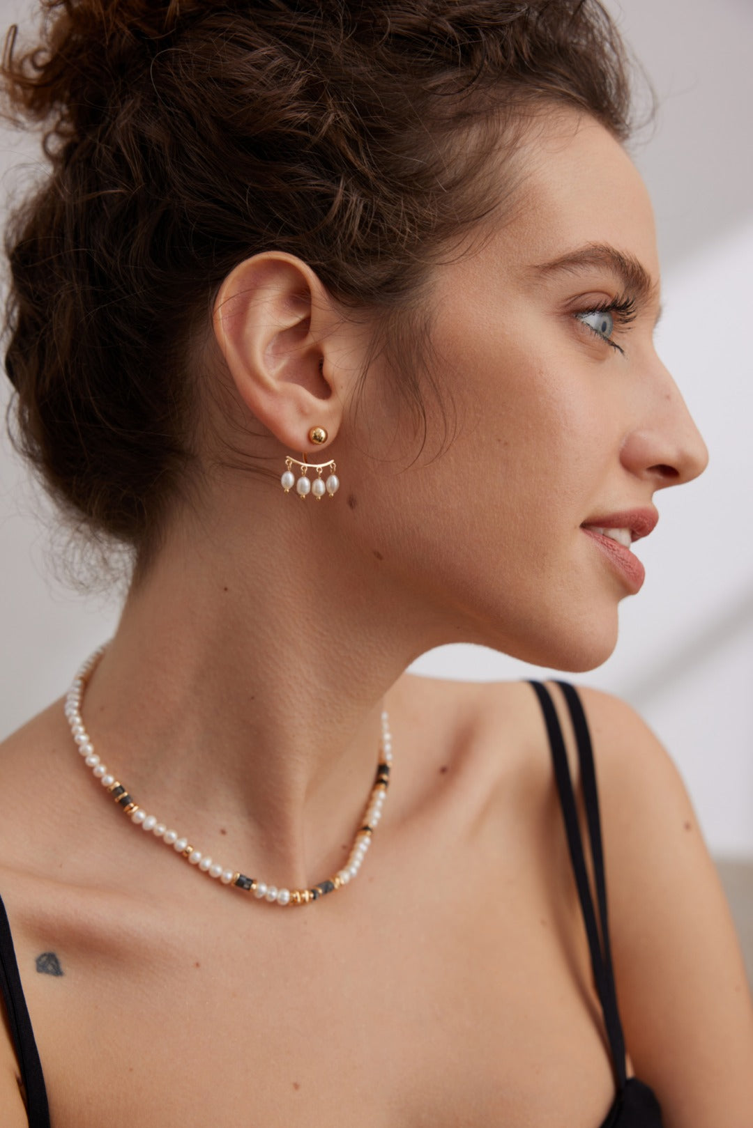 A woman with curly hair is facing to the right, wearing an elegant pair of Siren Mode's Vintage Gold 925 Silver Natural Pearl Drop Earrings along with a matching pearl necklace adorned with gold and black beads. She is dressed in a black sleeveless top against a neutral background.