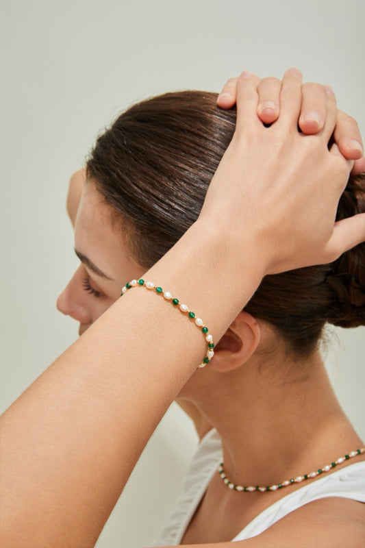 A person with dark hair styled in a bun is seen from the side, holding their hands on top of their head. They are wearing a green and white beaded bracelet paired with the Siren Mode Vintage Gold 925 Silver Natural Pearl Synthetic Emerald Bracelet and a matching necklace made from natural pearls. They are dressed in a white sleeveless top.