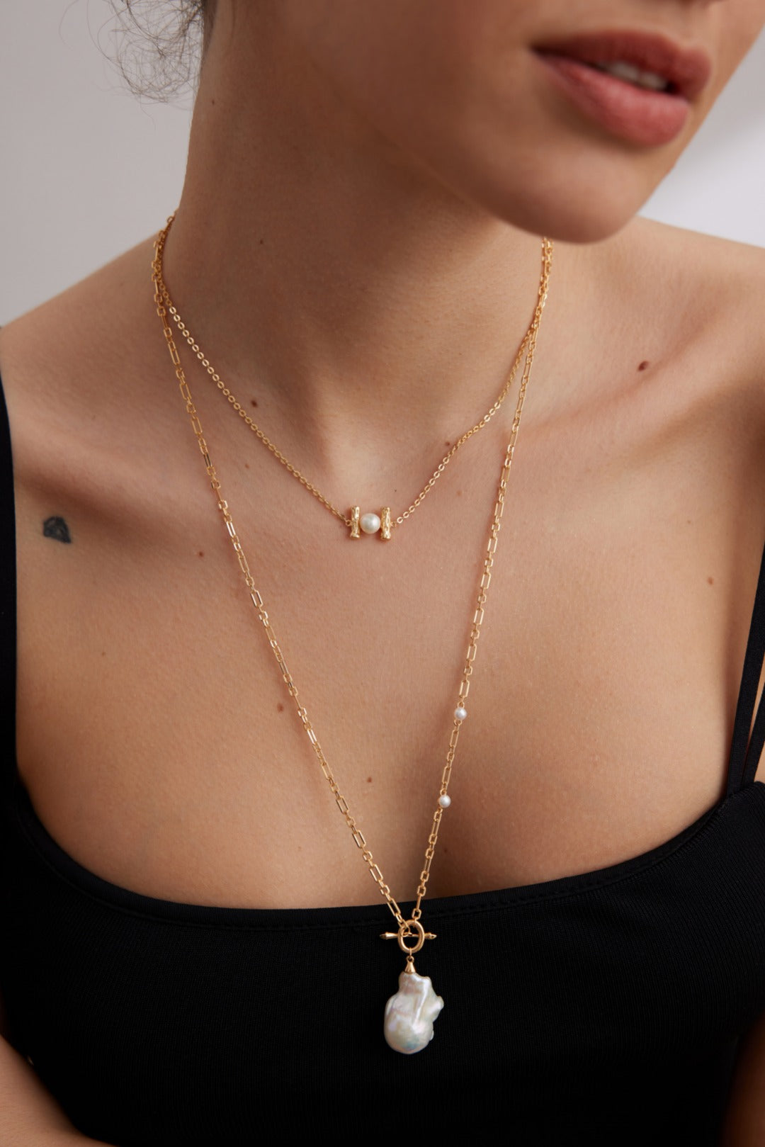 A person adorned in Siren Mode jewelry: they wear a stunning Vintage Gold 925 Silver Natural Pearl and Zircon Pendant Necklace with a shorter piece featuring small pearl embellishments and a longer chain showcasing an eye-catching large, irregular natural pearl pendant. The individual complements the jewelry with a black, sleeveless top and has a small tattoo on their shoulder.