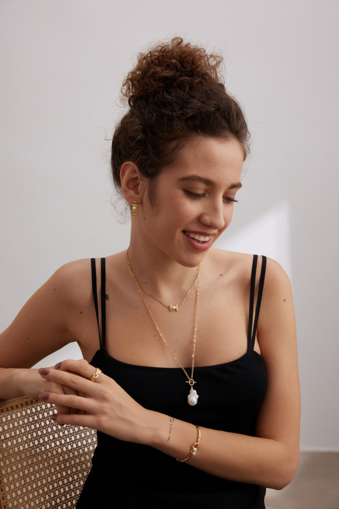 A woman with curly hair in a bun is wearing a black sleeveless top. Adorned with multiple pieces of gold jewelry, including earrings, necklaces, rings, and bracelets, she also sports the Siren Mode Vintage Gold 925 Silver Natural Pearl and Zircon Pendant Necklace. She is smiling and looking down to her side while seated on a woven chair.