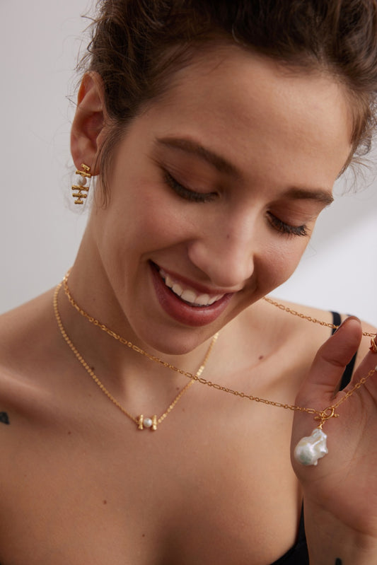 A woman smiles and looks down, wearing vintage gold jewelry from Siren Mode. She has on two necklaces and a pair of earrings. One necklace is the Siren Mode Vintage Gold 925 Silver Natural Pearl and Zircon Pendant Necklace, while the other features a small charm. With light makeup and her hair tied back, she appears to be in a well-lit indoor setting.