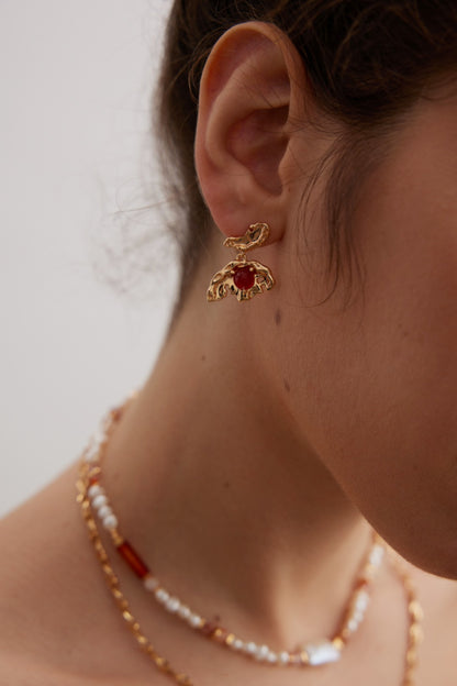 Close-up of a person's ear adorned with Siren Mode's Vintage Gold 925 Silver Red Agate Drop Earrings. The individual also wears layered necklaces with gold, white, and red beads. The background is neutral, focusing attention on the elegant jewelry.