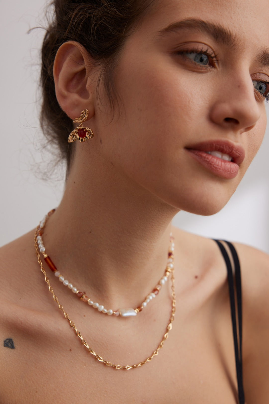 Close-up of a woman with light brown hair wearing gold and pearl jewelry, including Siren Mode's Vintage Gold 925 Silver Red Agate Drop Earrings, a layered necklace with pearls and small beads, and a thin gold chain necklace. She is posed against a neutral background and dressed in a black strap.