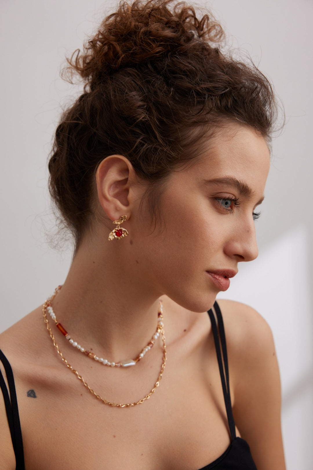 A woman with curly brown hair tied up in a bun is shown in profile. She is wearing Siren Mode's Vintage Gold 925 Silver Red Agate Drop Earrings, a layered necklace with beads and chain links, and a black spaghetti strap top. The background is out of focus and appears to be indoors.