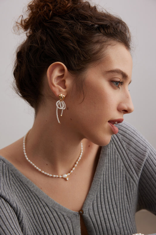 A woman with curly hair pulled into a bun is depicted in profile. She wears a ribbed, grey top, a 925 Silver necklace adorned with shell pearls, and Siren Mode's Vintage Gold 925 Silver Shell Pearl Drop Earrings. Set against a plain background, she looks slightly downward.