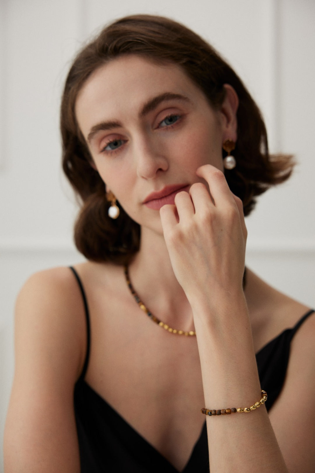 A person with short brown hair is wearing a black sleeveless top, pearl earrings, a beaded necklace, and the Vintage Gold 925 Silver Tiger's Eye Beaded Bracelet by Siren Mode. They are posing with their hand resting near their face, looking directly at the camera. The background is softly lit and blurred.