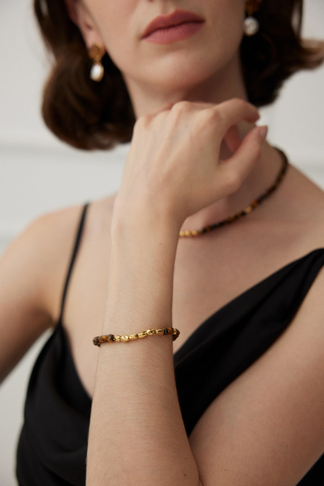 A person with short brown hair is wearing a black sleeveless top. They accessorize with gold and pearl earrings, a Siren Mode Vintage Gold 925 Silver Tiger's Eye Beaded Bracelet, a gold necklace, and a matching vintage gold bracelet. They are slightly turned to the side, with their left hand gracefully touching their neck.