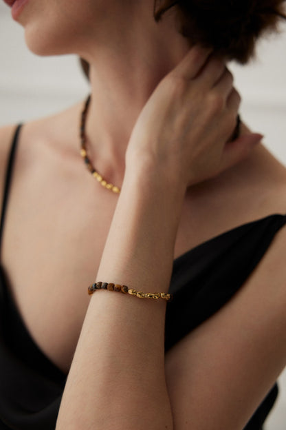 A woman in a black sleeveless dress is shown from the neck down, adorned with a matching stone and gold jewelry set. She rests her hand on her neck, prominently displaying the Siren Mode Vintage Gold 925 Silver Tiger's Eye Beaded Bracelet. The background is softly blurred.