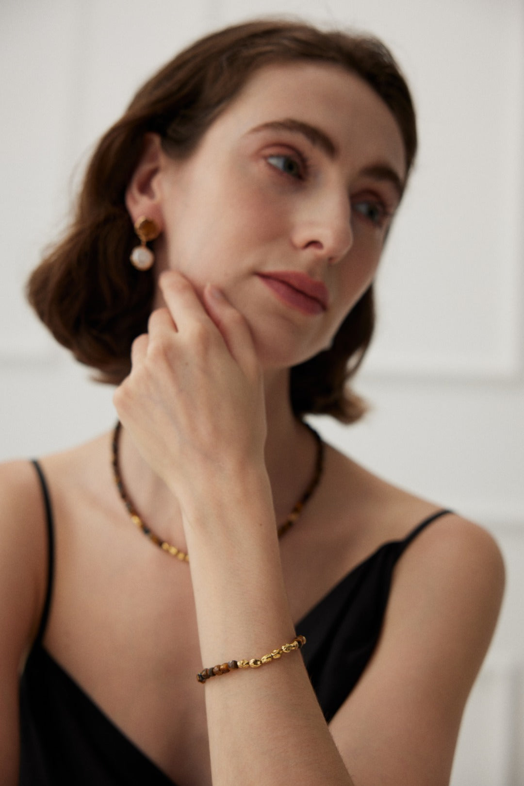 A woman with short brown hair is looking to the side, resting her chin on her hand. She is wearing a black sleeveless top and accessorized with gold jewelry, including a Siren Mode Vintage Gold 925 Silver Tiger's Eye Beaded Bracelet, along with a vintage gold necklace and earrings featuring pearl details. The background is softly blurred.