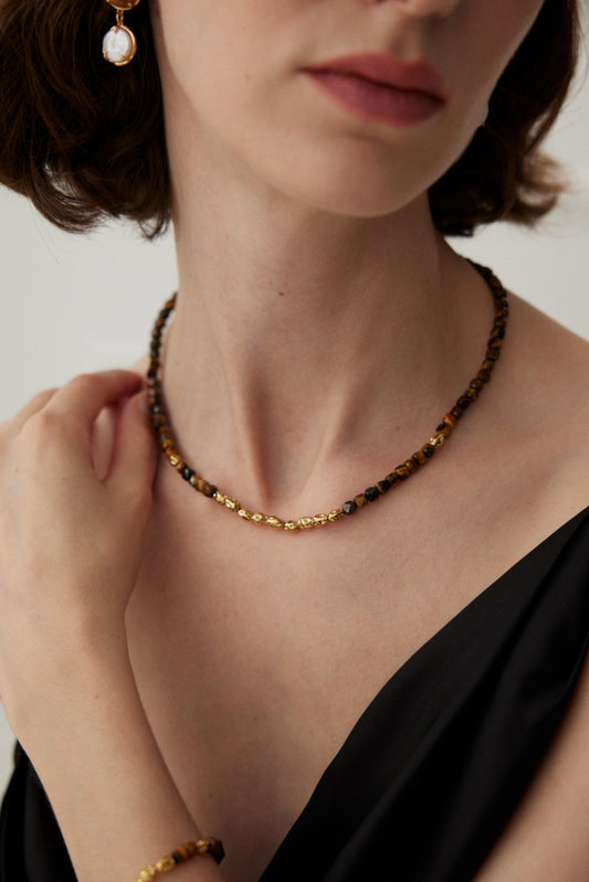 Close-up of a person wearing a dark, sleeveless top. They are adorned with a delicate Siren Mode Vintage Gold 925 Silver Tiger's Eye Beaded Necklace and matching bracelet. They are touching the necklace with one hand against a plain, light-colored background.