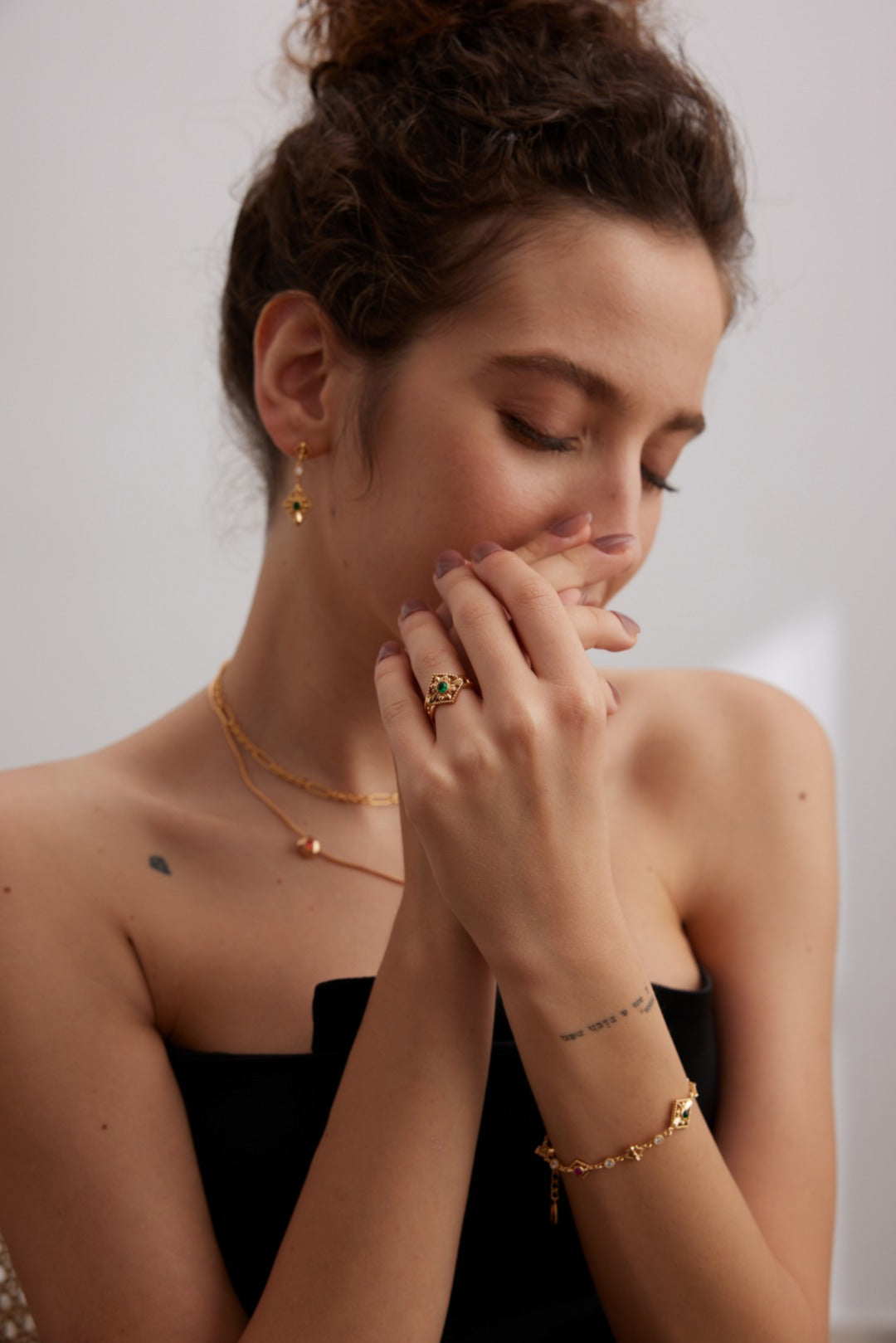 A woman with curly hair stands against a minimalist background, adorned in gold jewelry. She showcases her Vintage Gold 925 Silver Zircon Bracelet by Siren Mode along with zircon stone earrings, a necklace, and several rings. Wearing a black strapless top, she poses with her eyes closed and hands near her face to highlight the elegance of the jewelry.