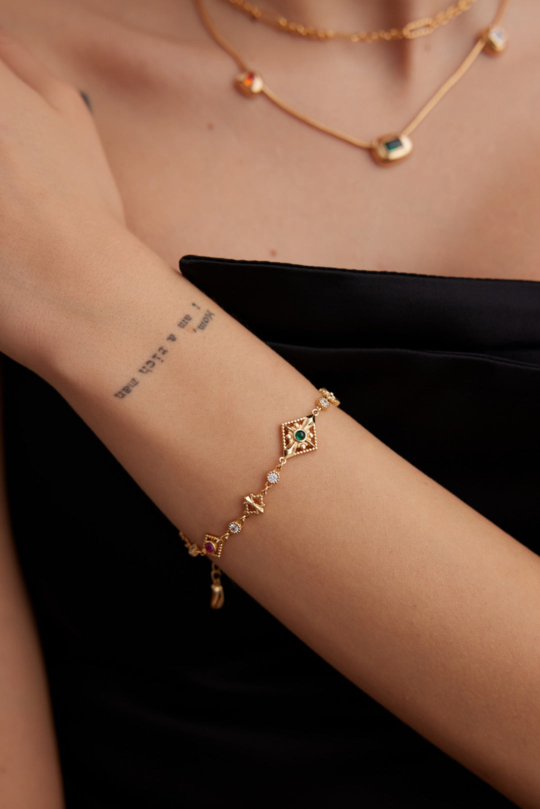 A close-up of a person's arm adorned with Siren Mode's Vintage Gold 925 Silver Zircon Bracelet. The person is also wearing a layered gold necklace with geometric pendants. There's a tattoo with text on their forearm, and the background is kept neutral.