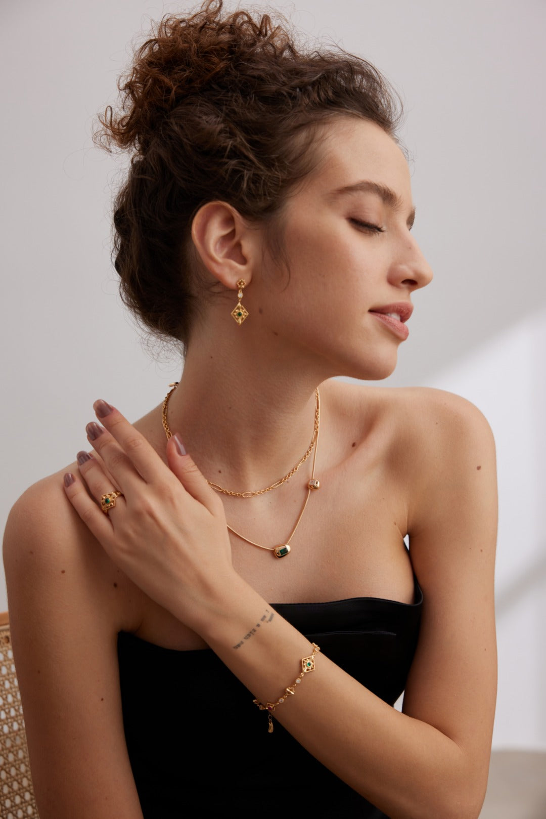 A woman with curly hair in a bun is posing with her eyes closed, adorned in gold jewelry featuring earrings, layered necklaces, a ring, and the Vintage Gold 925 Silver Zircon Bracelet by Siren Mode. She wears a strapless black top and exudes a relaxed, serene expression.