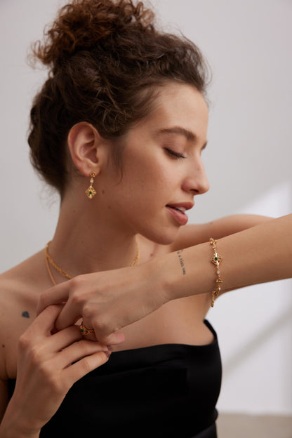 A woman with curly hair is wearing a black off-shoulder top and various gold jewelry, including necklaces, earrings, and a Vintage Gold 925 Silver Zircon Bracelet from Siren Mode. She is looking down at her hand to showcase the bracelet adorned with dazzling zircon stones. The background is softly lit and neutral.