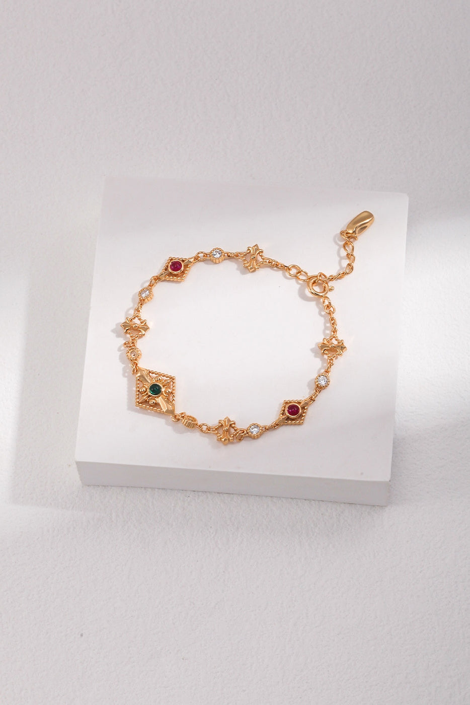 A delicate Siren Mode Vintage Gold 925 Silver Zircon Bracelet featuring intricate designs and colorful gemstone embellishments, placed on a white square display against a softly lit background. The chain includes small, intricate patterns with red and green zircon gems interspersed along its length, reminiscent of timeless elegance.